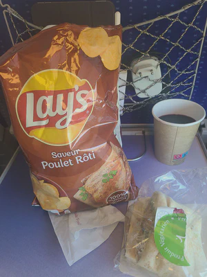 Picture of a folded down tray at a seat on a train. On the tray is a bag of roast chicken flavored potato chips (Lays 'Saveur poulet rôti' flavor), a half-eaten vegetarian sandwich in cellophane, paper cup of red wine, and a napkin.