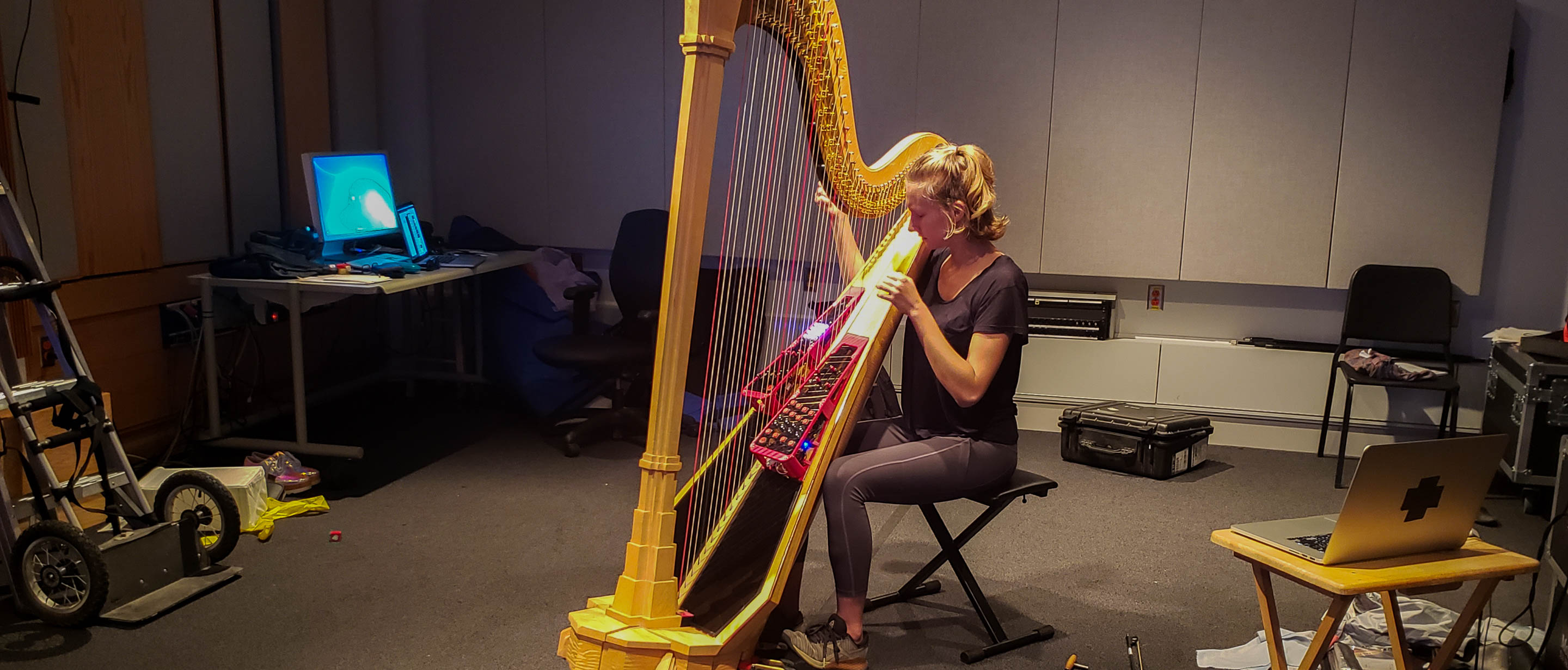 A woman sitting in the middle of a large but messy research studio. She is playing a concert harp that is equipped with two electronic controllers that are fitted on either side of the harp strings. They rise up at an angle from the soundboard, and each has a layout of several knobs, buttons, and sliders. The units are made of red 3D printed frames and black acrylic panels. Around the room is strewn a large amount of research equipment: computers, black cases, a violin bow, and other miscellaneous objects.