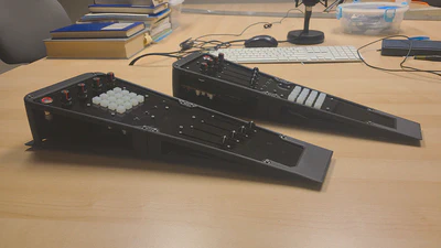 Two hardware controllers sit on a desk. They are made out of black #D printed frames and black acrylic panels, and feature layouts of buttons, knobs and sliders.