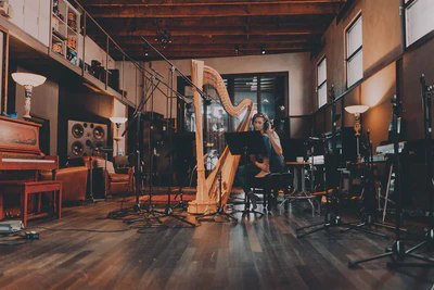 A photo of a woman in a recording studio sitting next to a harp. She is listening to something back in the headphones. The studio is cozy and full of wood, and there are pianos, amplifiers, and other instruments lining the walls.