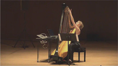 A woman seated on stage playing a concert harp. A computer sits on a table next to her and a music stand is in front of her. Visible on the back of the performer's hand is a small device, which is the wireless gesture acquisition device.