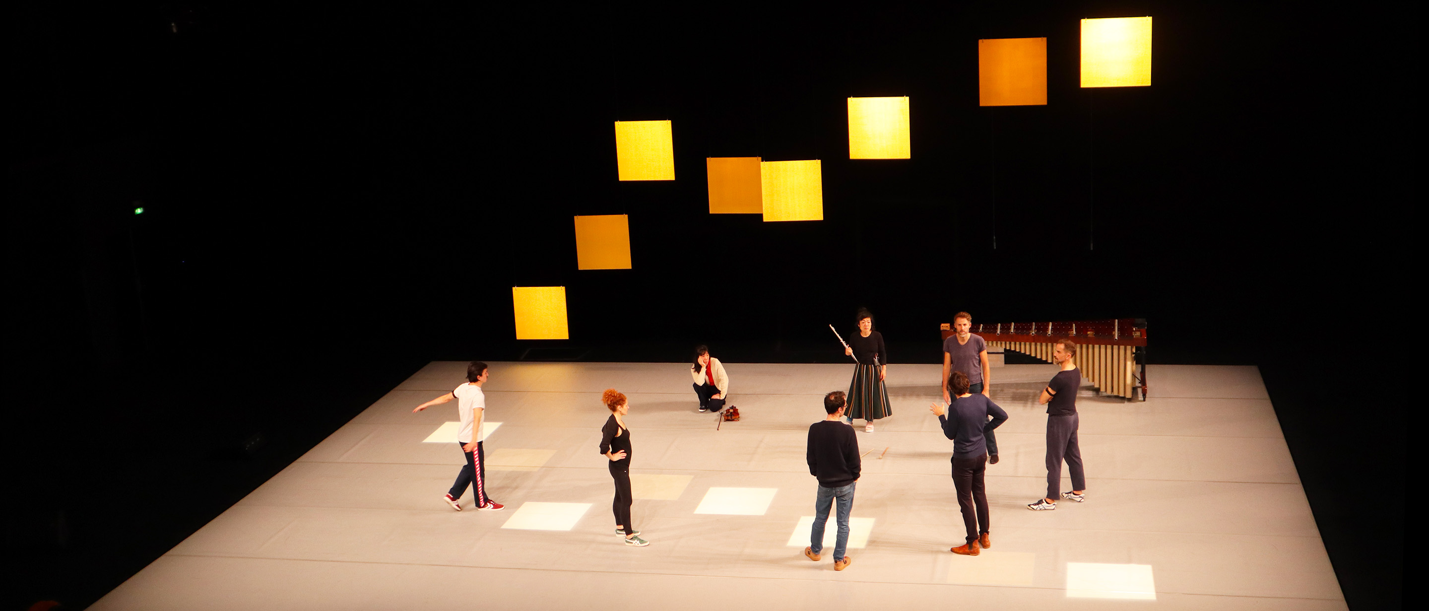 A photograph of a stage in a theater. The walls are black and the floor is white. illuminated by the overhead stage lights. Eight square panels hang at the back of the stage, some light up, some not. The same squares are also projected on the floor of the stage in the same pattern. A marimba sits in the back right corner of the stage. Eight individuals are in a circle in the middle of the stage, in discussion during a break in the rehearsal.