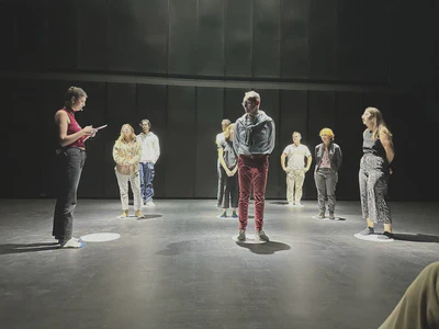 Nine people stand on a stage, forming a 3 by 3 grid. Each stands on a white circle on the ground and are bathed in white light from spotlights above. One person is reading from a paper while the others watch.