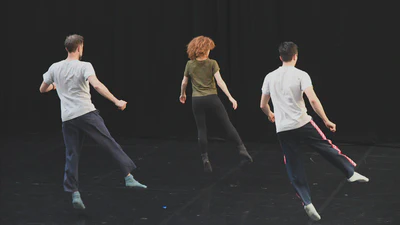 Three dancers execute a synchronized move. The are in a line with their backs to the camera and in mid-air, with their right leg slightly kicked out to the side.