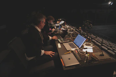 Looking down the long table of the control booth at the back of the theater. A man sit sitting in front of two computers, and a woman sits beyond him working at a sound board.
